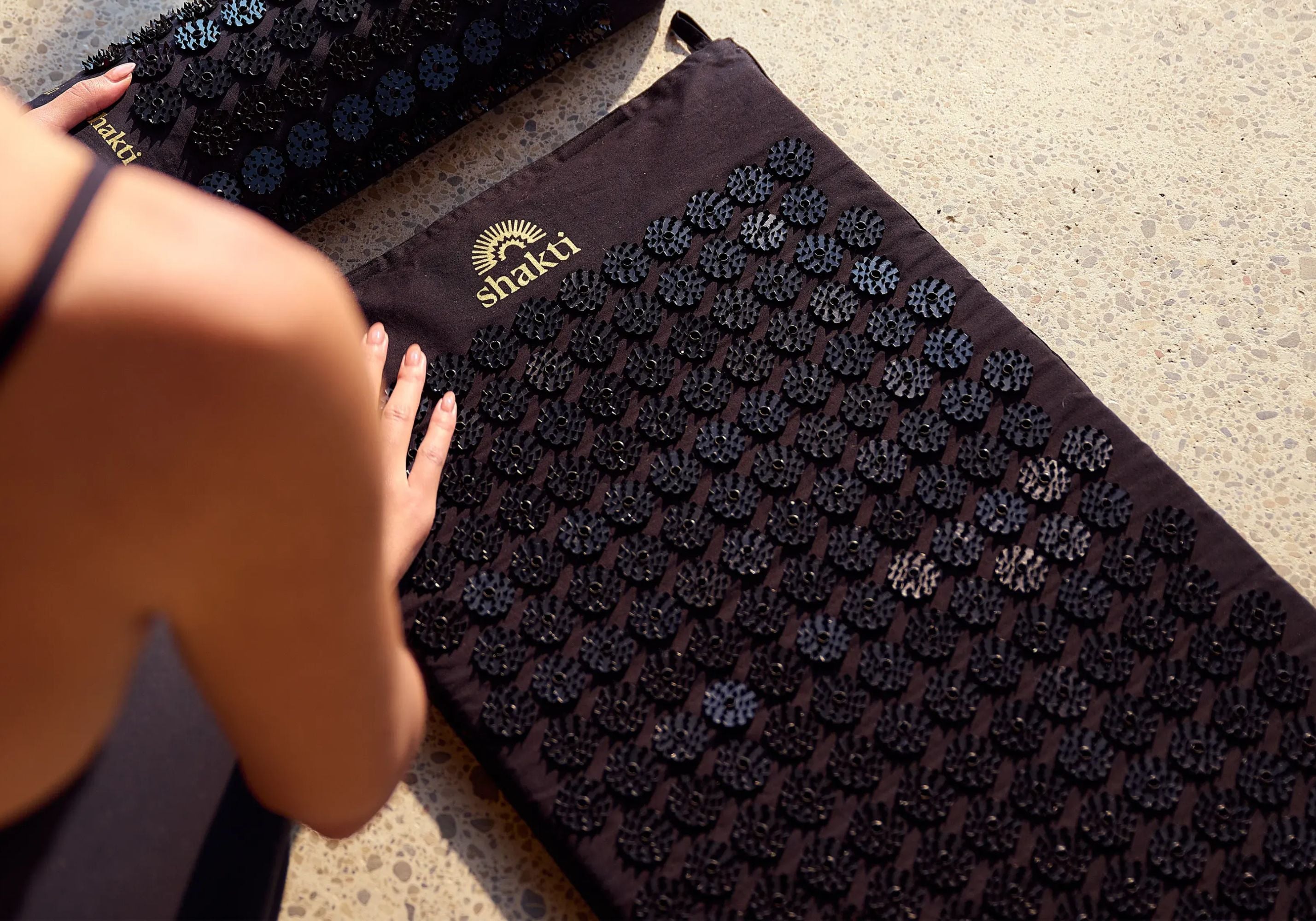 A person laying a Shakti Classic Mat in Black onto a textured concrete floor.