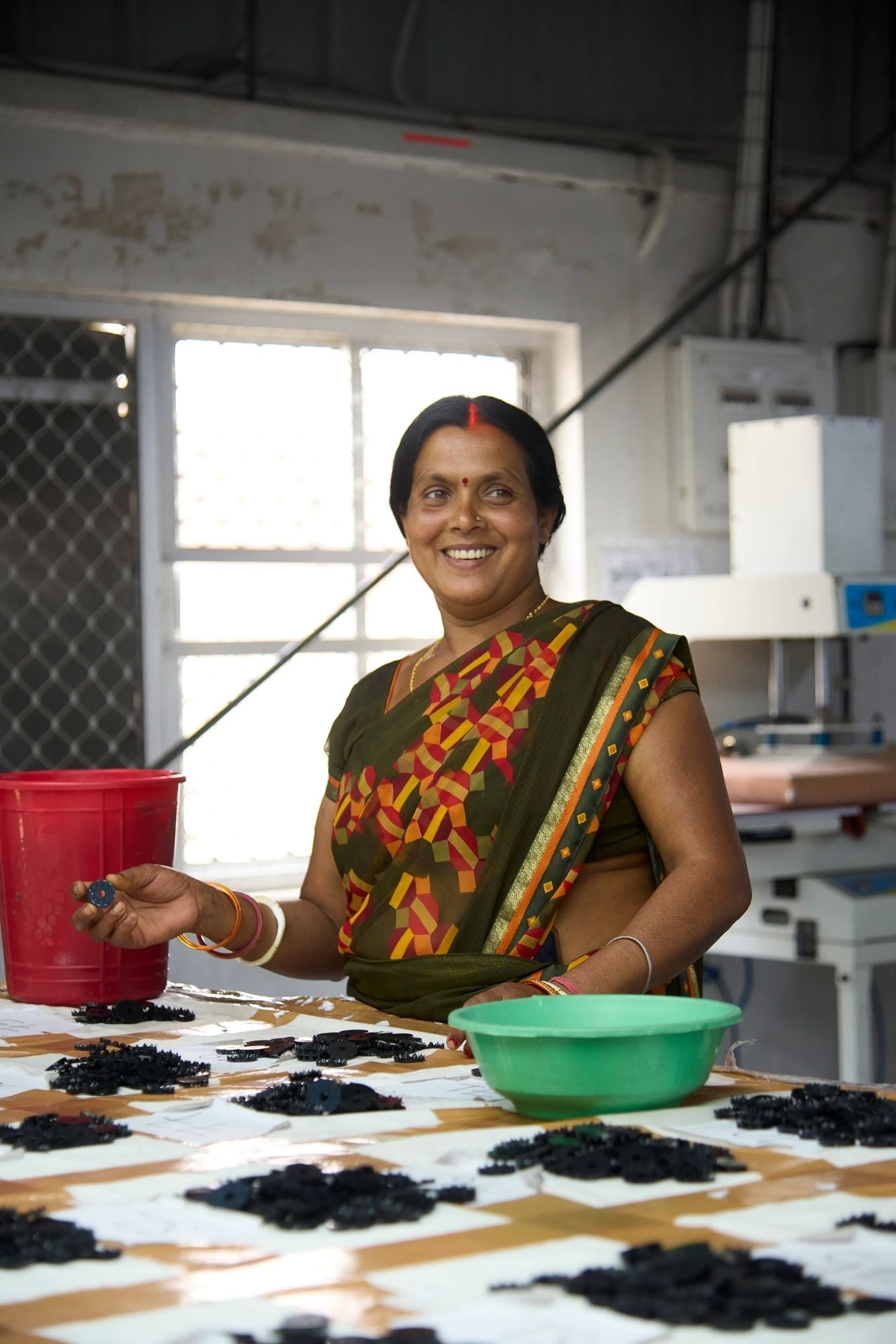Woman smiling in a colorful saree sorting signature Shakti spikes an acu-disks in a factory setting.